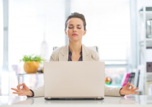 Image of a woman sitting in front of a laptop, her eyes closed and fingertips touching in a meditation pose