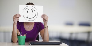 Person sitting at desk holding a piece of paper with a happy face drawn onto it